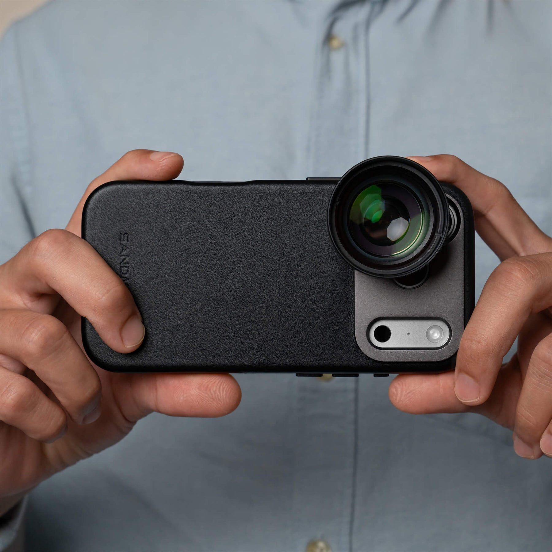 Two hands holding an iPhone 18 Pro Max in a SANDMARC Black Pro Leather Case horizontally with the Telephoto 2x Lens attached, worn by a man in a light blue shirt showing the SANDMARC logo and multi-coated green glass element
