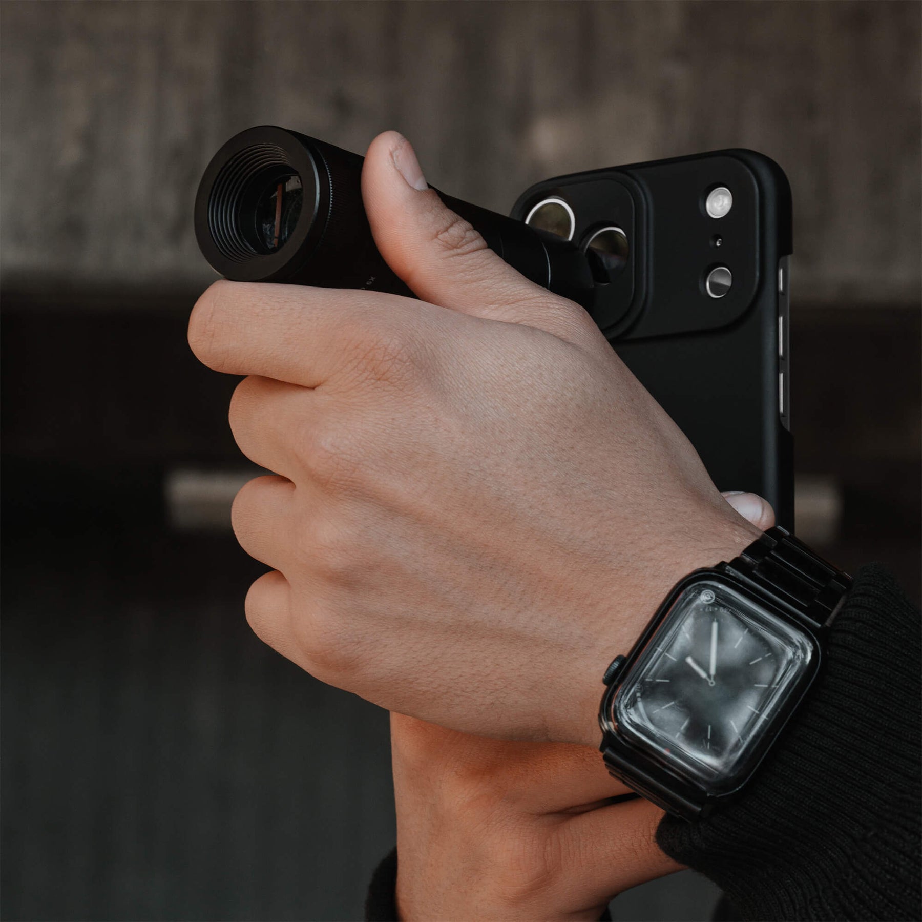 Hand gripping an iPhone 18 Pro Max in a SANDMARC Black Standard Case with the Telephoto 6x Lens extended forward, wearing a SANDMARC Titanium Apple Watch Band against a blurred dark indoor background