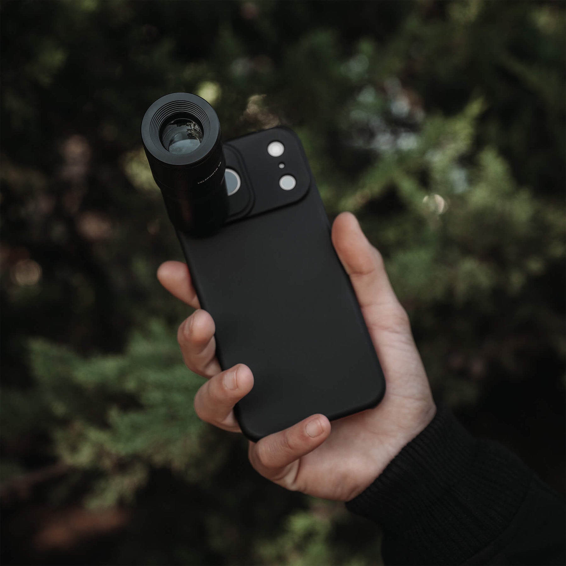 Hand holding an iPhone 18 Pro Max in a SANDMARC Black Standard Case with the Telephoto 6x Lens attached, held up against a blurred dark green pine tree background