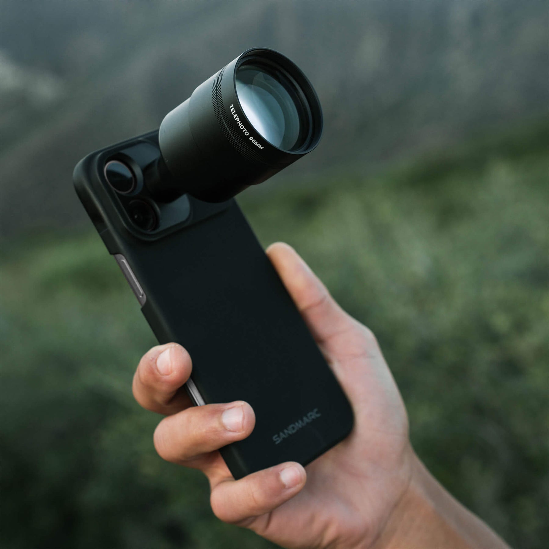 Hand holding an iPhone 18 Pro Max in a SANDMARC Black Standard Case with the Telephoto Tetraprism 96mm Lens attached, showing the Telephoto 96MM barrel label, held outdoors against a blurred dark green mountain landscape