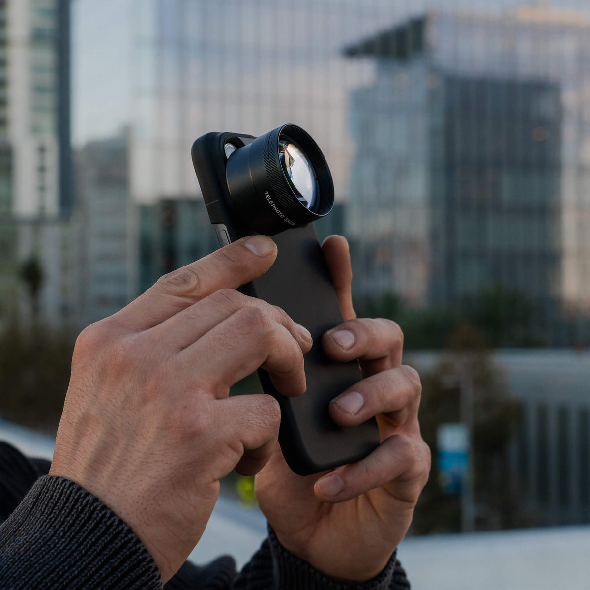 Two hands adjusting the SANDMARC Telephoto 2x Lens attached to an iPhone 18 Pro Max in a SANDMARC Black Standard Case outdoors, with blurred glass office buildings visible in the background at dusk