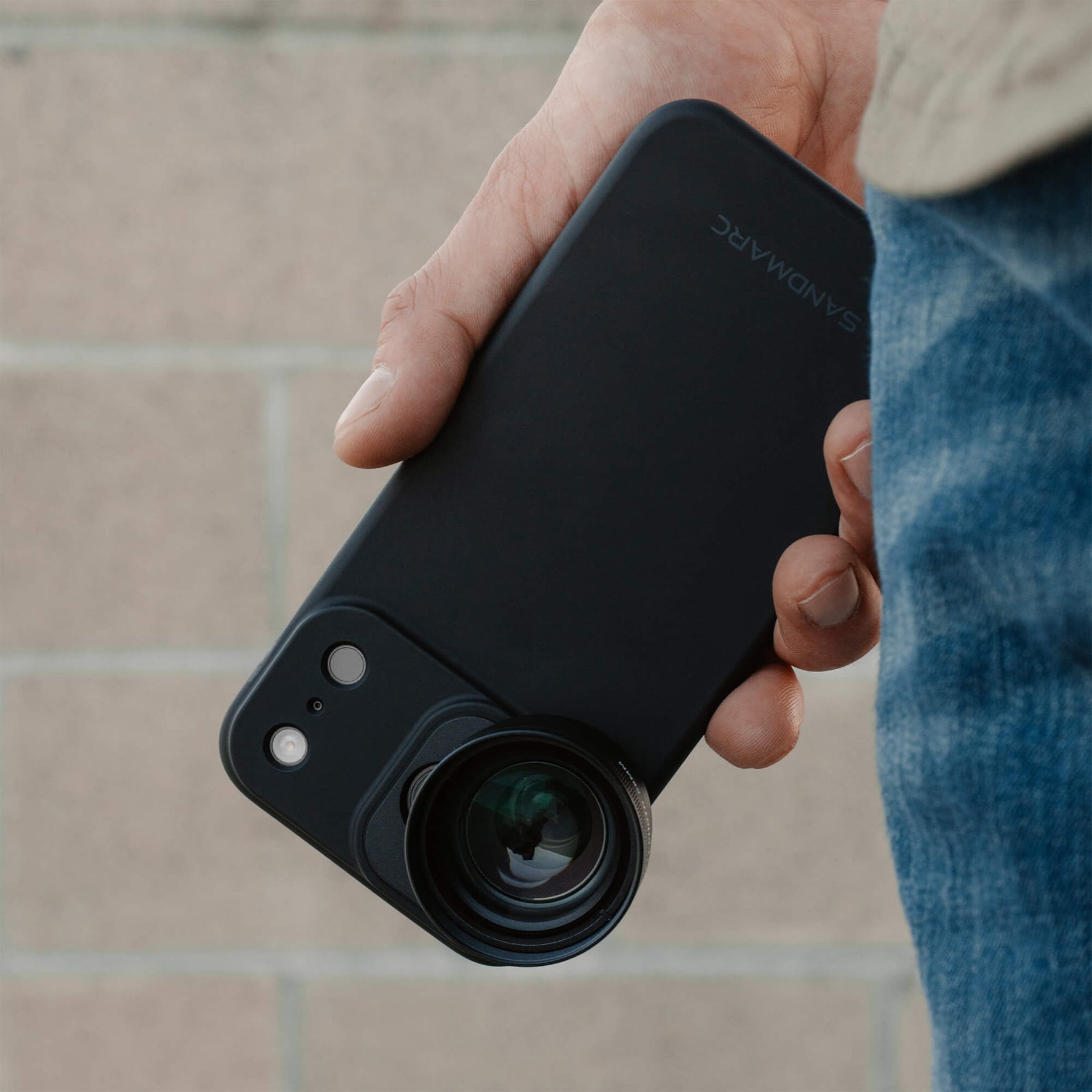 Hand holding an iPhone 18 Pro Max in a SANDMARC Black Standard Case at his side with the Telephoto 2x Lens attached at the bottom, wearing blue jeans against a beige brick wall background