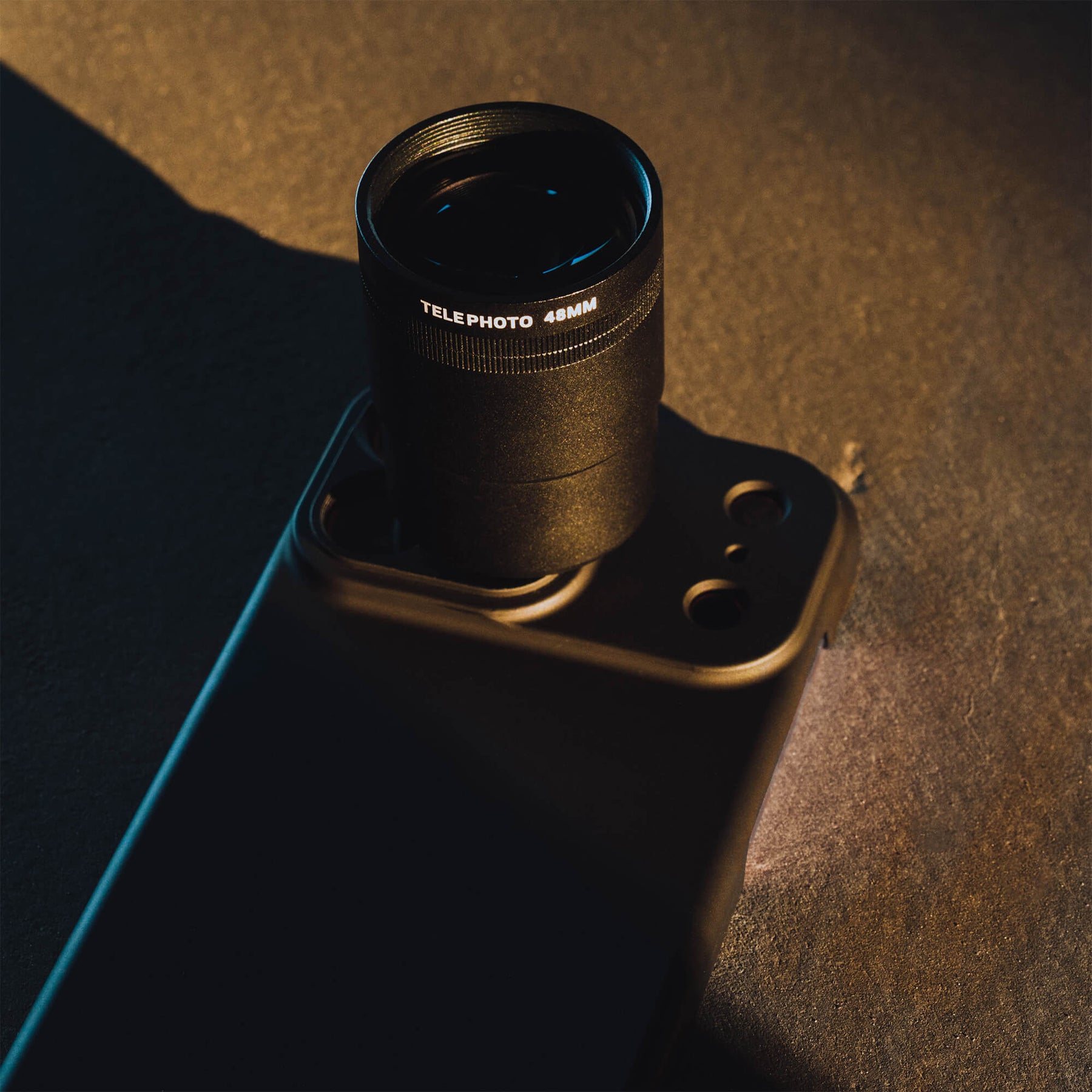 SANDMARC Telephoto Tetraprism 48mm Lens mounted on an iPhone 18 Pro Max in a SANDMARC Black Standard Case resting on a dark rust-textured concrete surface in dramatic golden hour sidelight, showing the Telephoto 48MM barrel label and lens detail