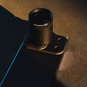 SANDMARC Telephoto Tetraprism 48mm Lens mounted on an iPhone 18 Pro Max in a SANDMARC Black Standard Case resting on a dark rust-textured concrete surface in dramatic golden hour sidelight, showing the Telephoto 48MM barrel label and lens detail