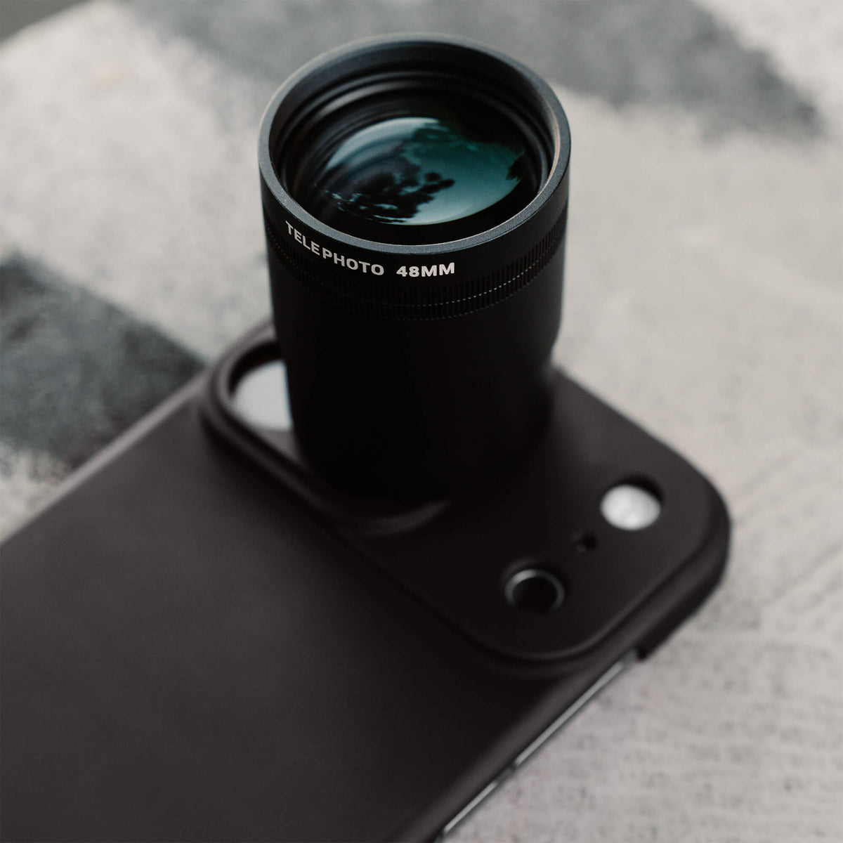 SANDMARC Telephoto Tetraprism 48mm Lens mounted on an iPhone 18 Pro Max in a SANDMARC Black Standard Case, resting on a grey and white textured fabric surface, showing the Telephoto 48MM barrel label and multi-coated teal glass element