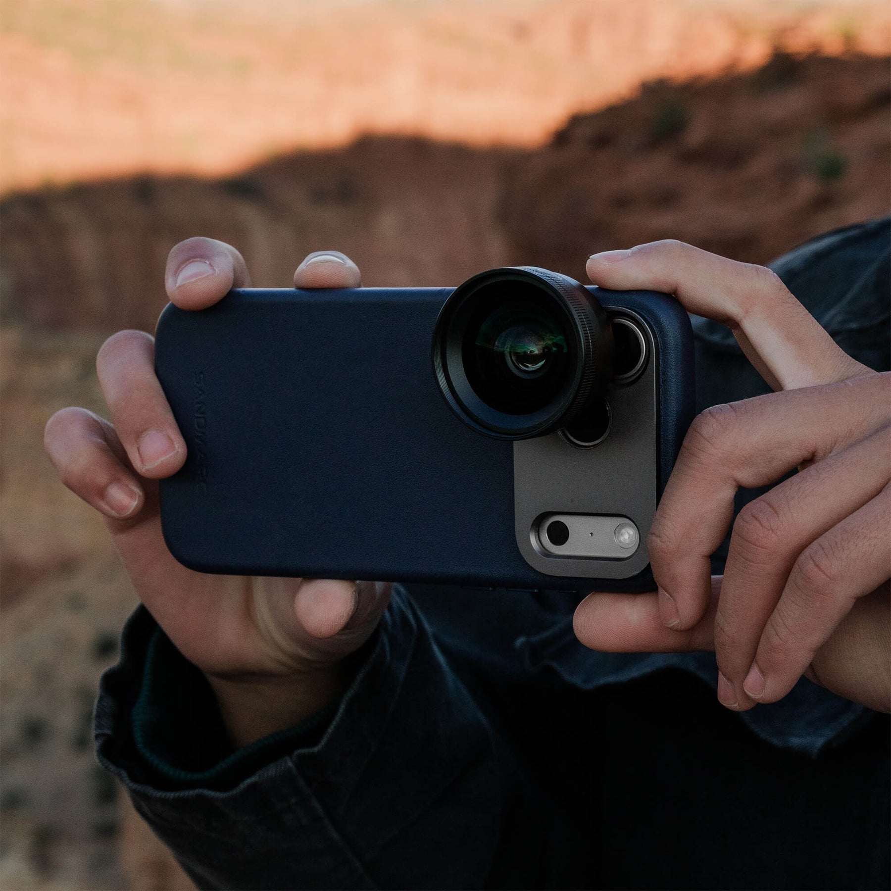Person holding an iPhone 18 Pro Max in a SANDMARC Navy Pro Leather Case with the Wide Lens 16mm attached horizontally at golden hour, with red canyon cliffs visible in the background