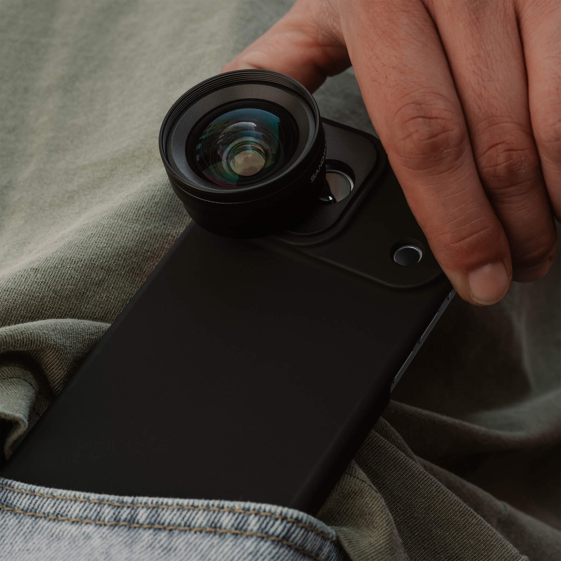 Close-up of a hand pulling an iPhone in a SANDMARC Black Standard Case with the Wide Lens 16mm attached from a pocket, with sage green shirt and denim jean pocket visible in the background