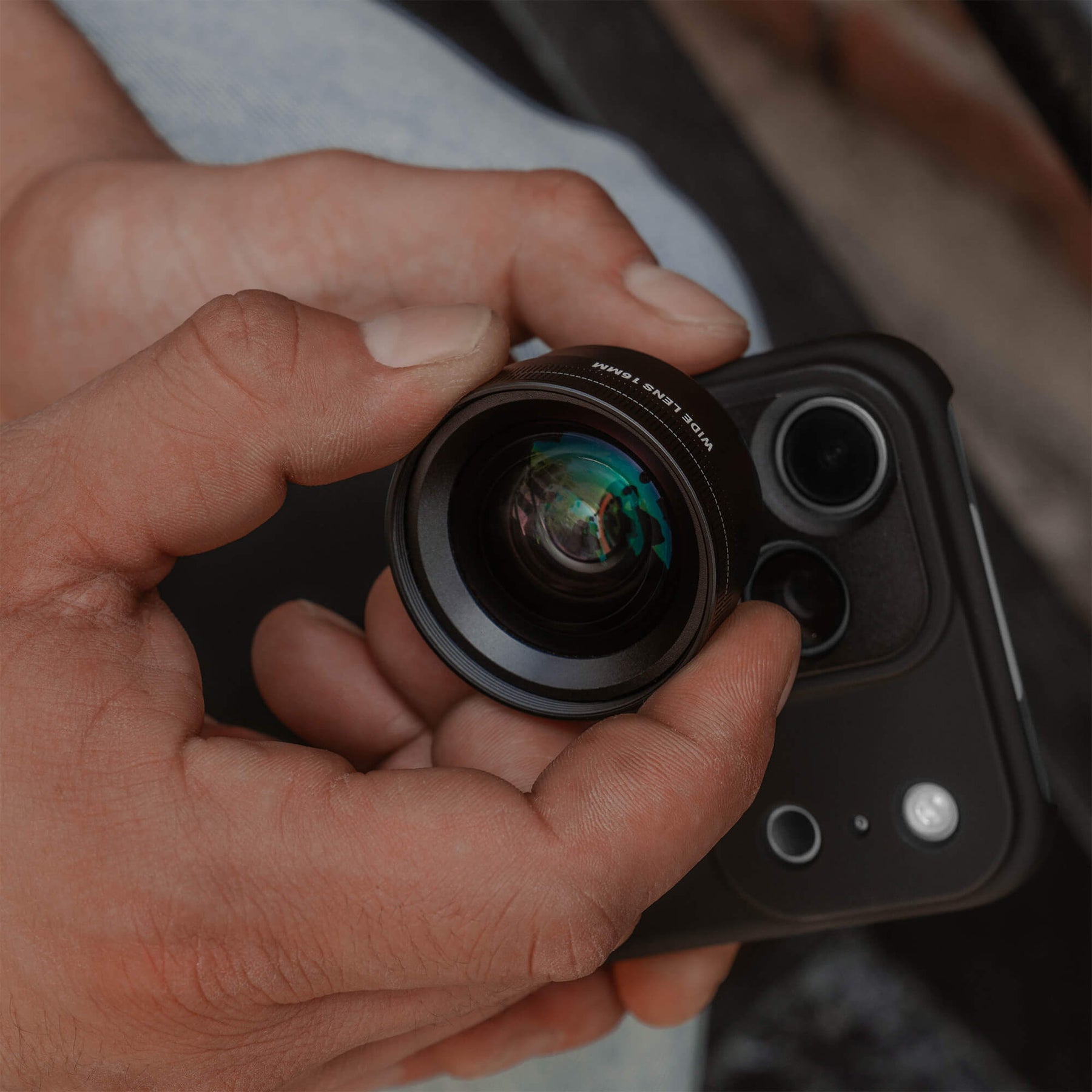 Close-up of a hand holding the SANDMARC Wide Lens 16mm in front of an iPhone in a SANDMARC Black Standard Case, showing the multi-coated glass elements and lens barrel detail