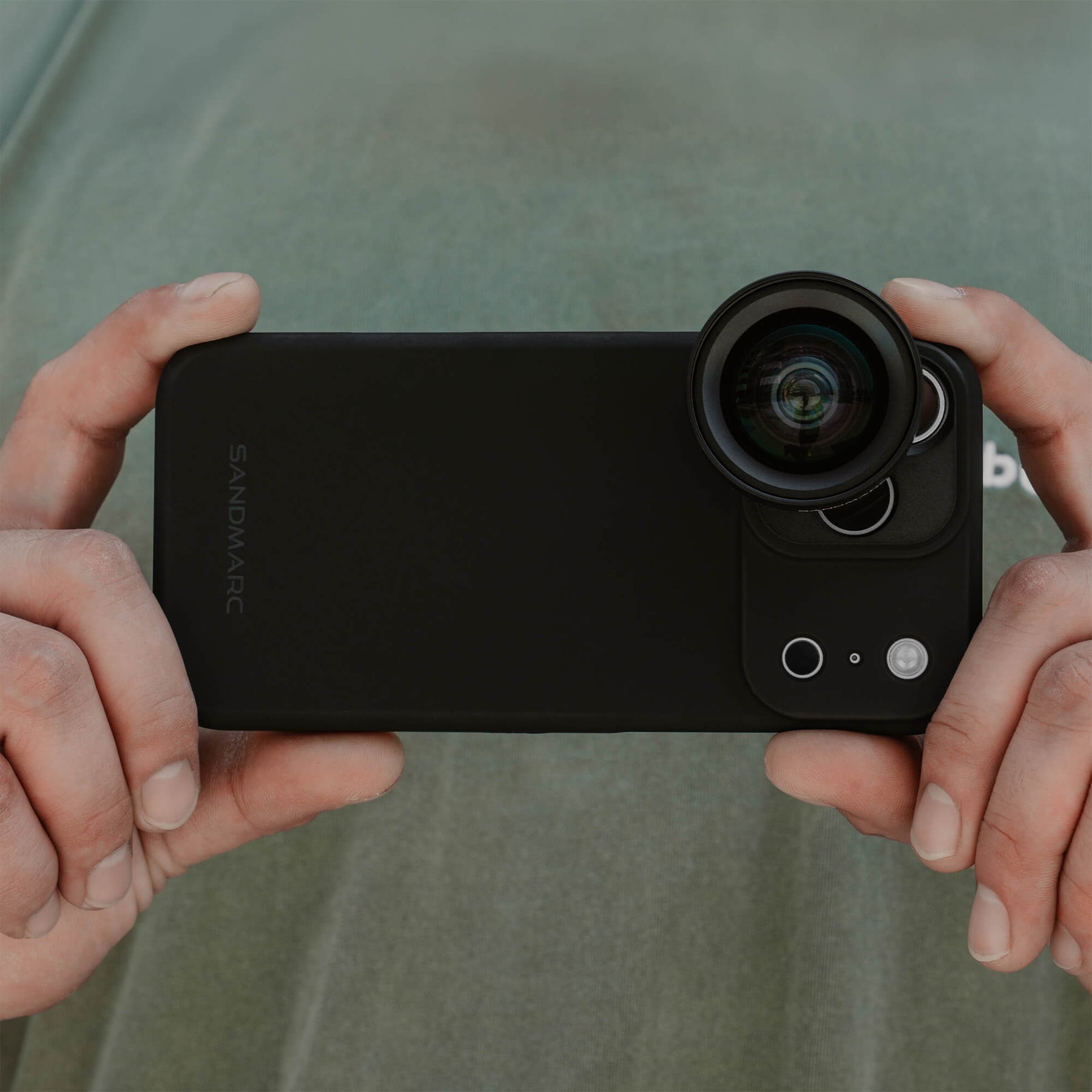 Two hands holding an iPhone 18 Pro Max in a SANDMARC Black Standard Case horizontally with the Wide Lens 16mm attached, photographed against a sage green shirt